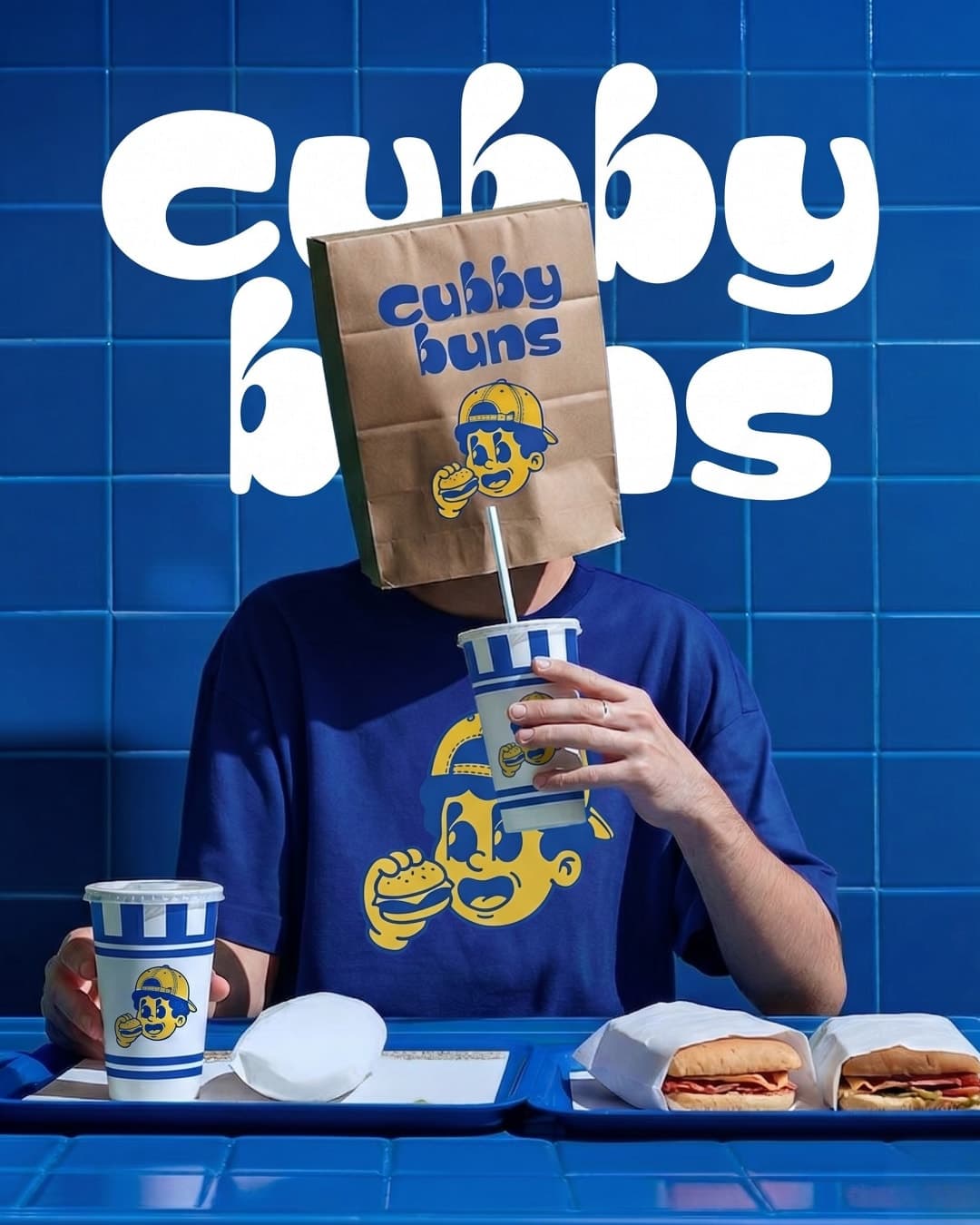 Person with a Cubby Buns paper bag over their head, sipping a branded cup at a blue-tiled counter