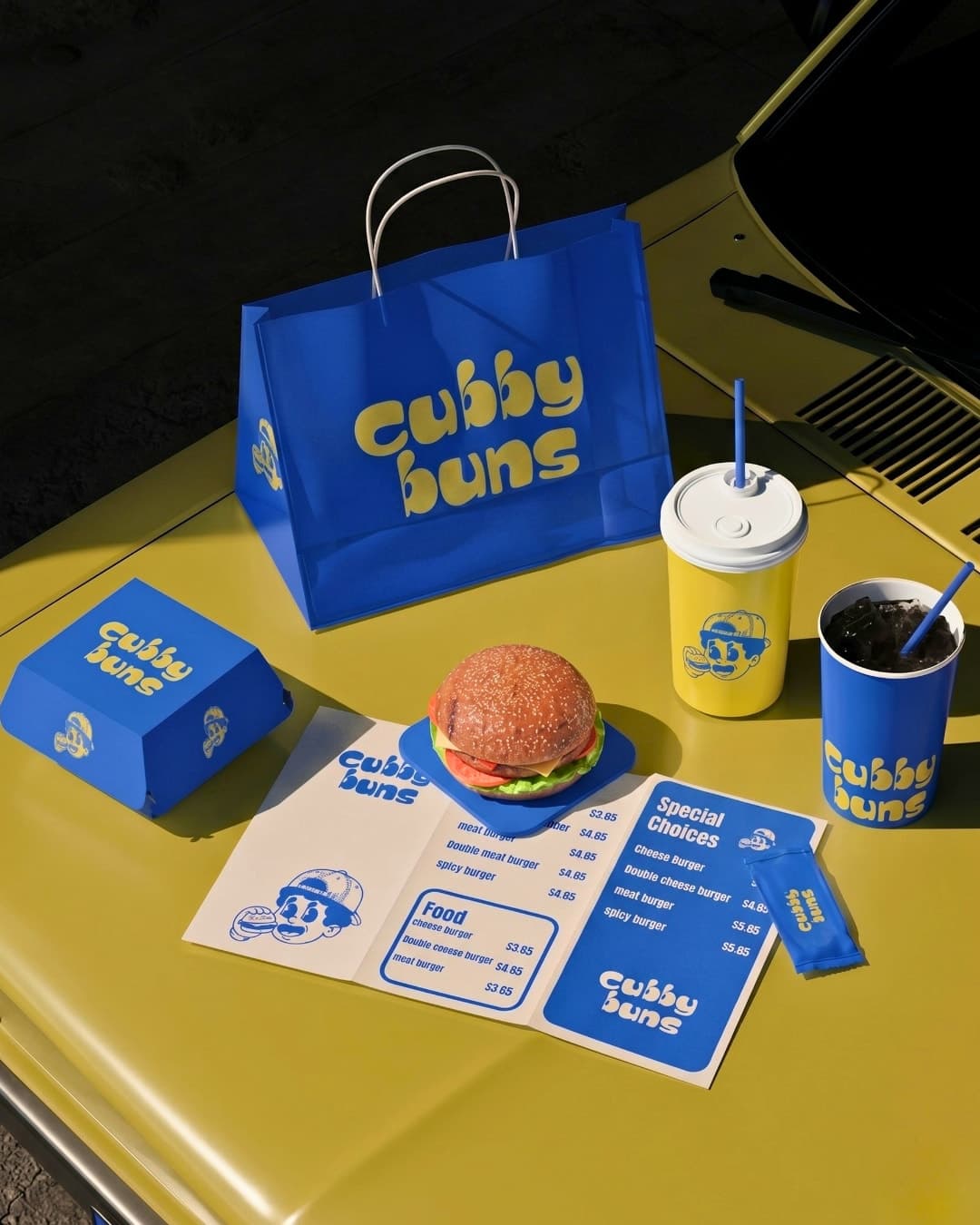 Full Cubby Buns packaging set — bag, cup, box, burger and menu — on a yellow car bonnet
