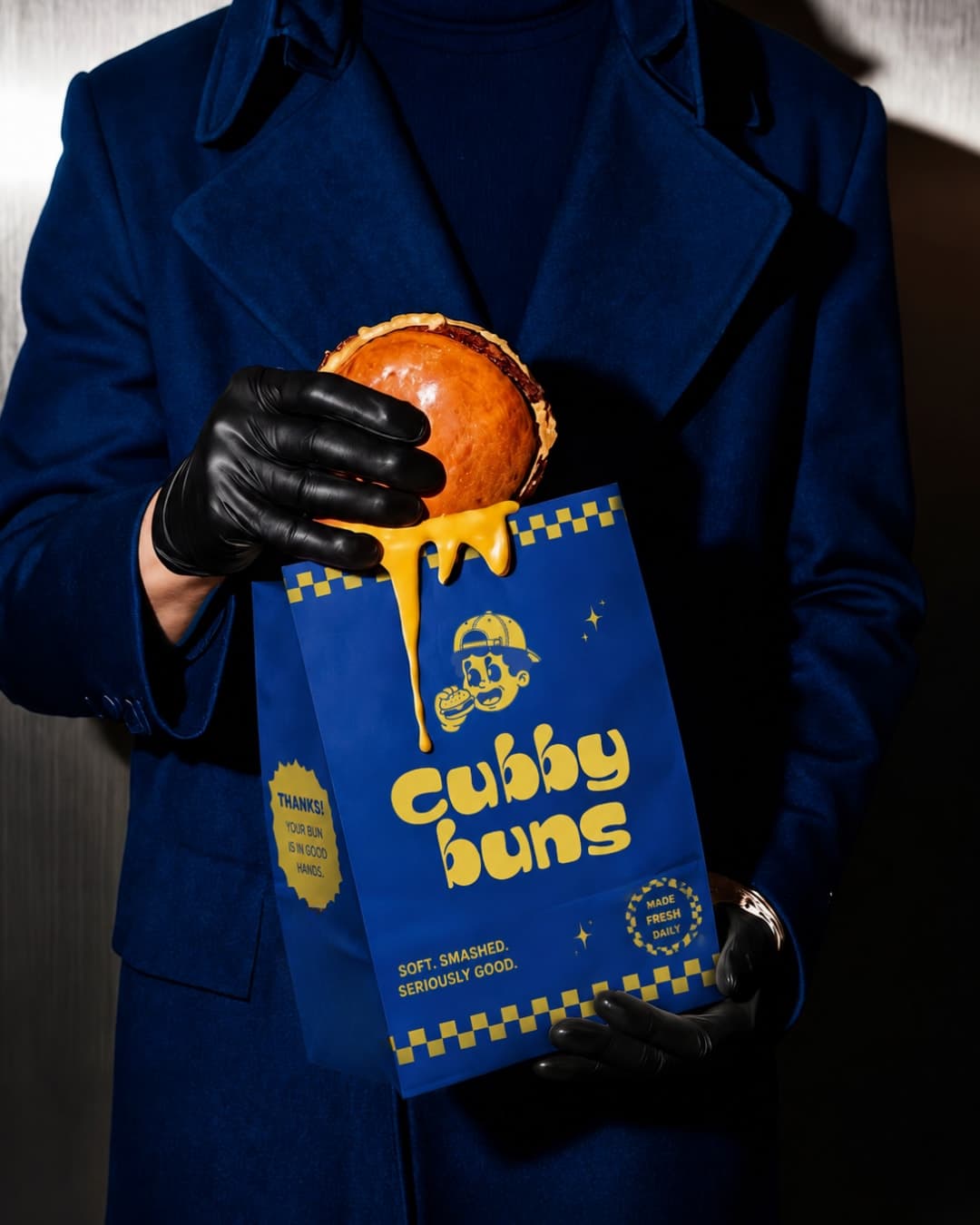 Gloved hand holding a dripping burger over a blue Cubby Buns bag
