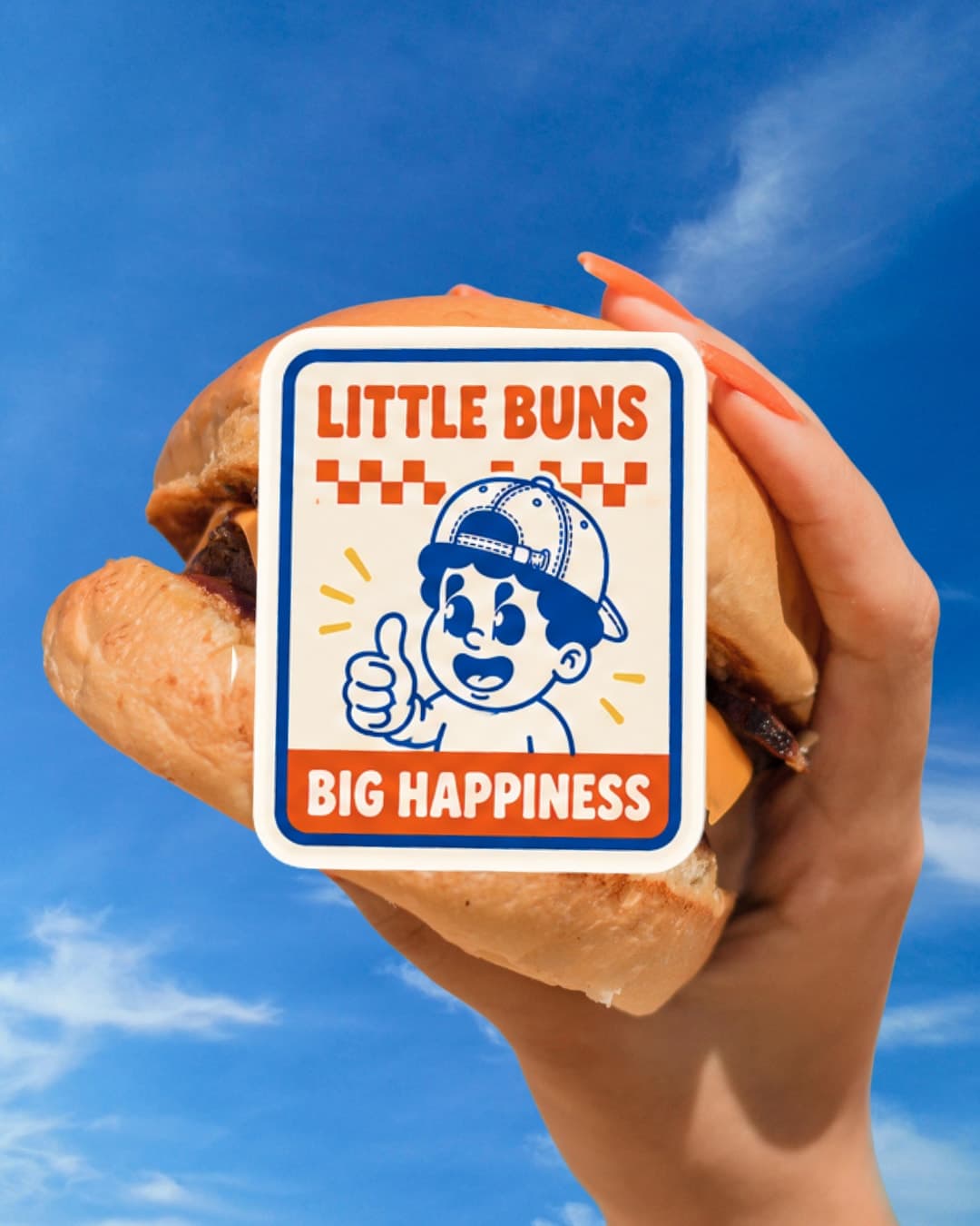Hand holding a burger with the 'Little Buns, Big Happiness' sticker against a blue sky